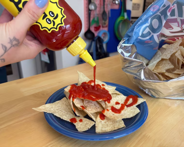 A hand squeezing a bottle of Yellowbird Classic Jalapeño Hot Sauce, adding some to a tortilla chip.