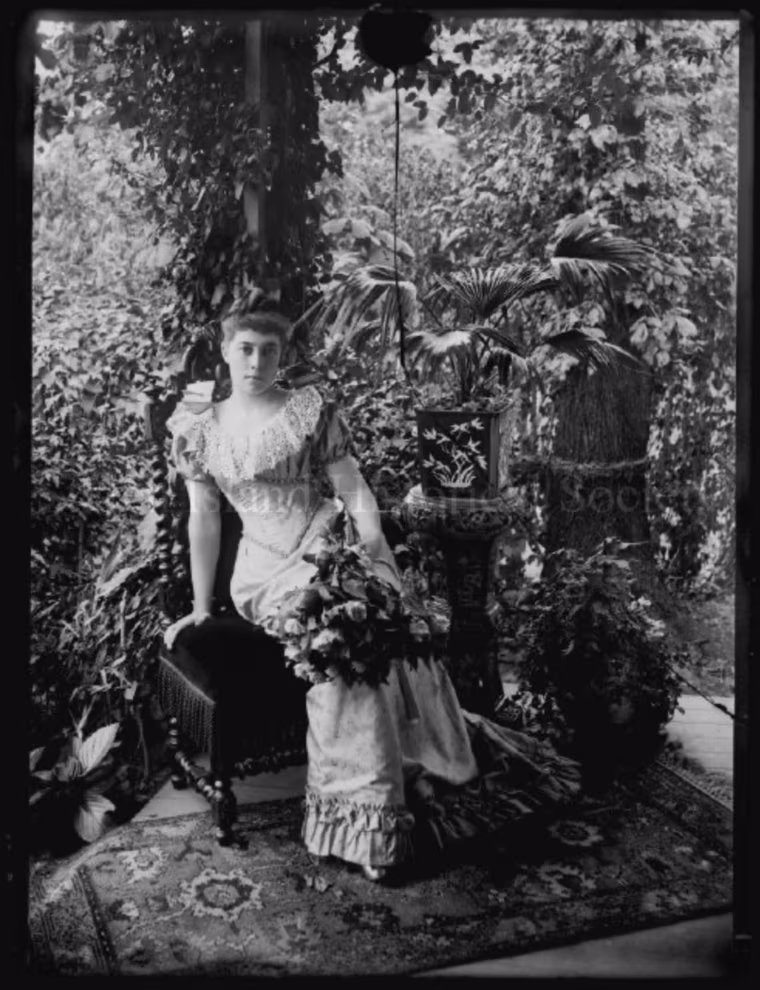 Full length, with bouquet,
self-portrait by Alice Austen, front porch of Clear Comfort, 1892.