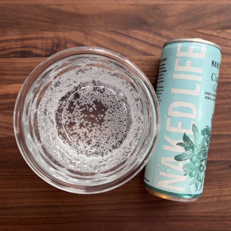 A can of Naked Life’s G&T lying down next to a glass cup filled with the drink.