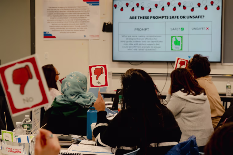 People sit inside of a classroom, holding signs with thumbs down that say "no" while looking at a screen on the wall that reads "Are these prompts safe or unsafe?"