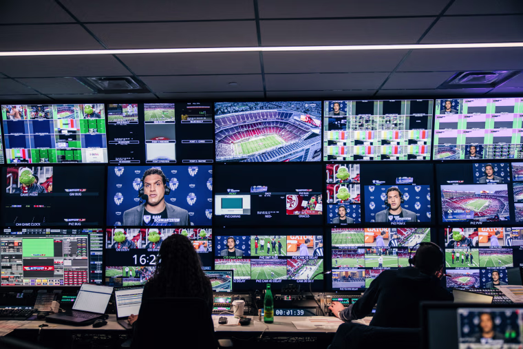 People sit in a control room in front of many computer and television screens displaying various angles of a football game
