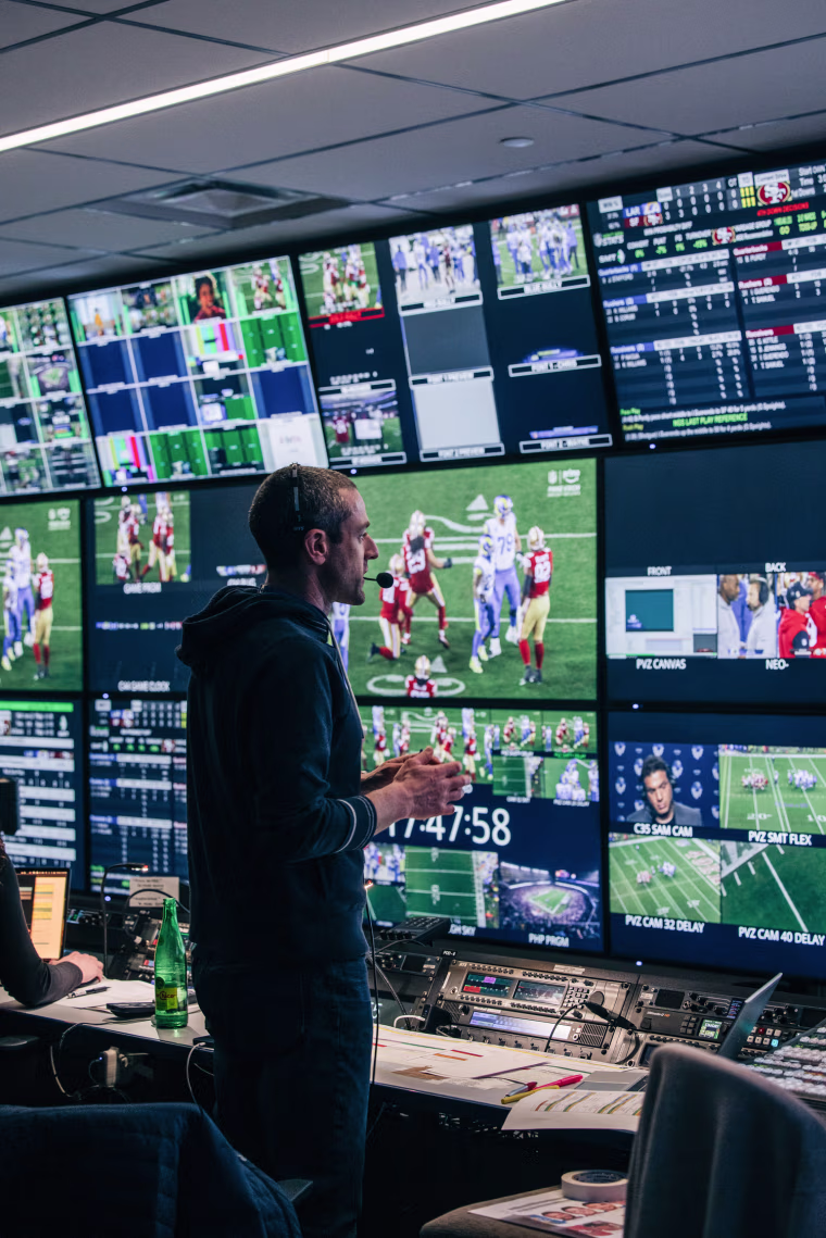 A person inside of a control room looks at various television screens stacked atop one another displaying different angles of a football game