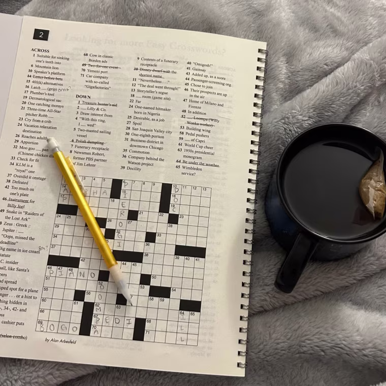A picture of a crossword puzzle with a pencil, next to a cup of tea, on a grey blanket.