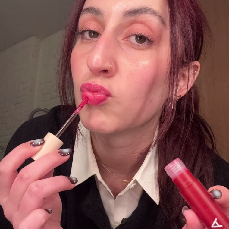 Woman applying red lip gloss to puckered lips while holding a gloss tube.