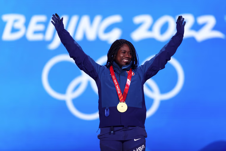 Erin Jackson raises her arms in celebration, wearing a gold medal necklace in front of a blue backdrop with white text that reads: Beijing 2022"