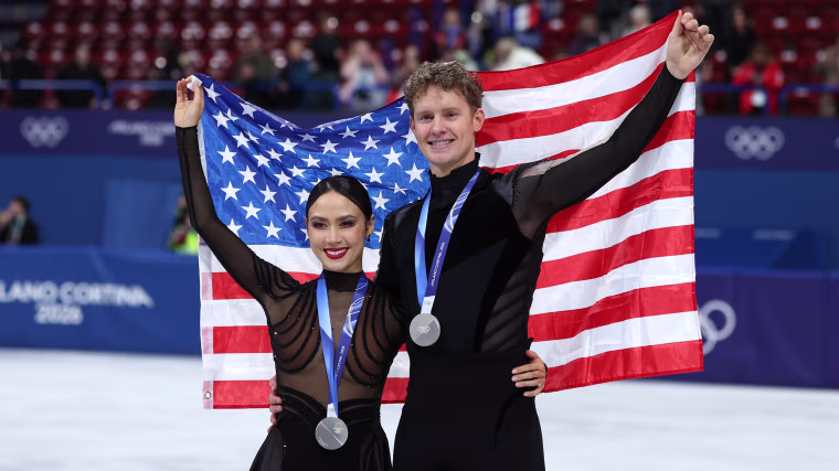 Madison Chock and Evan Bates hold an American flag up behind them while wearing silver Olympic medals around their necks