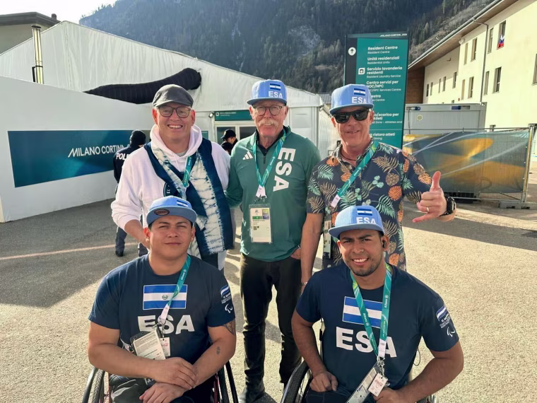 Three men stand behind two other men in wheelchairs outside a large white tent, with mountains behind them.