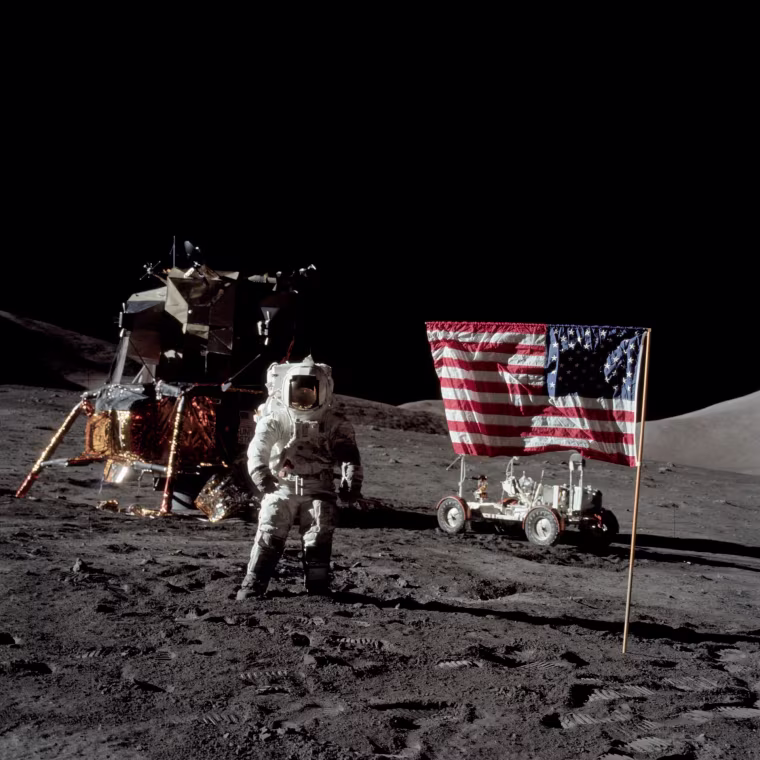 Harrison Schmitt stands on the surface of the moon in a spacesuit, with an American flag posted in the ground on his right, and a lunar module pilot on his left