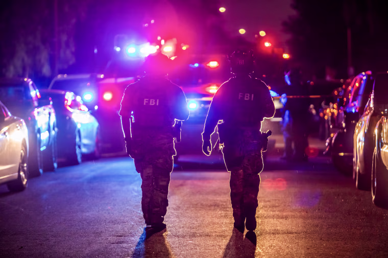 Two FBI field agents wearing helmets and camouflage fatigues walk in front of a vehicle with red and blue flashing lights.