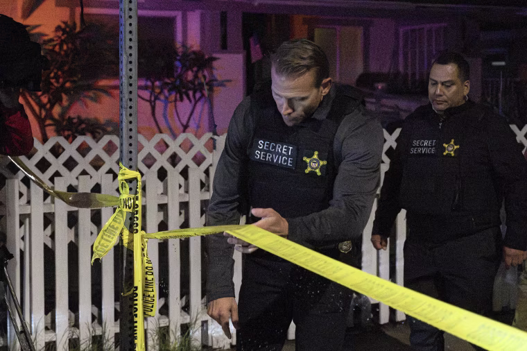 Two field agents wearing black flak jackets with Secret Service patches on them cross a yellow police perimeter.