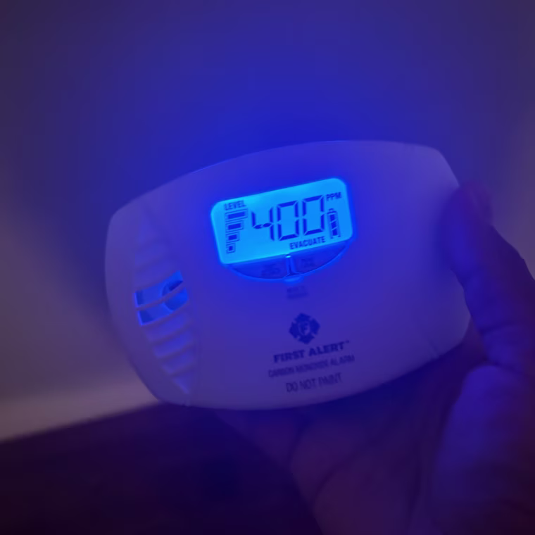 Person holding First Alert CO615 Dual-Power Plug-In Carbon Monoxide Detector with right hand in the dark and the blue backlit screen reading “Evacuate” and “400.”