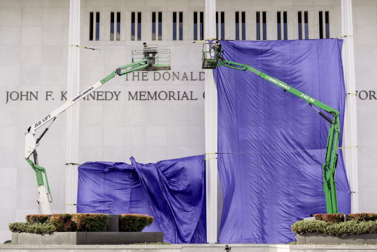 Workers in two hydraulic cranes are suspended above the ground in front of the Kennedy Center's facade, with a giant purple tarp stretched behind them.