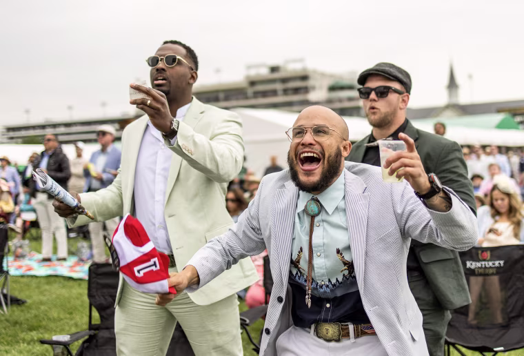 Three men cheer while standing in the infield of Churchill Downs under gray skies.