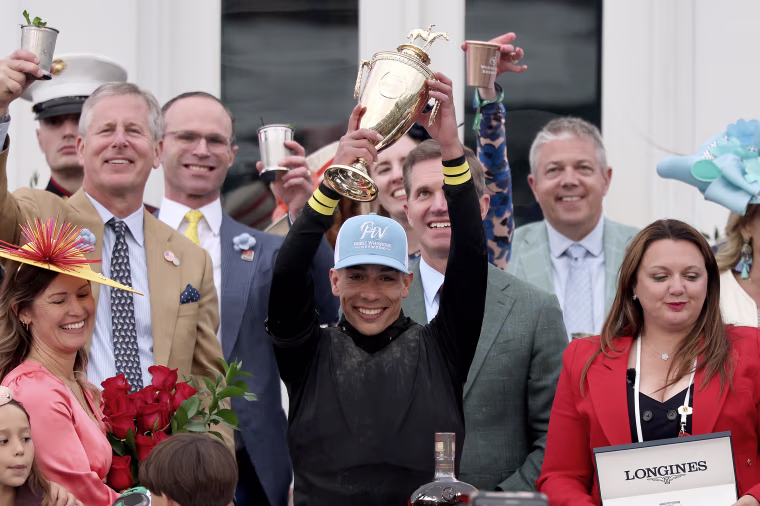 A jockey holds up a trophy while smiling broadly, surrounded by other celebrants.