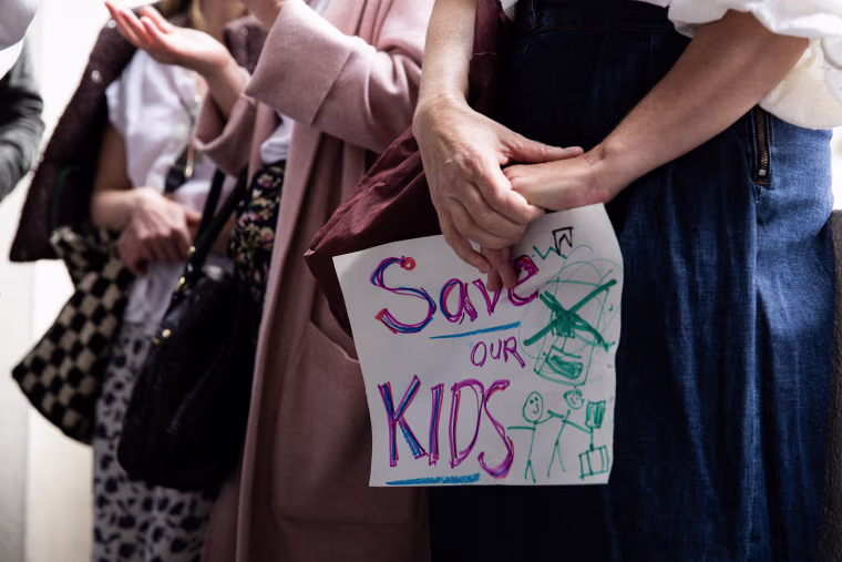 A person seen from the waist down holding a hand-written sign reading Save Our Kids.
