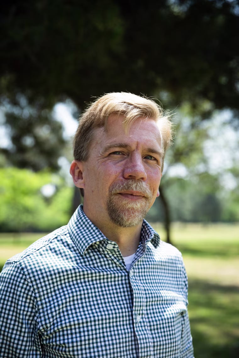 A goateed man wearing a checkered shirt standing for a portrait in the shade.