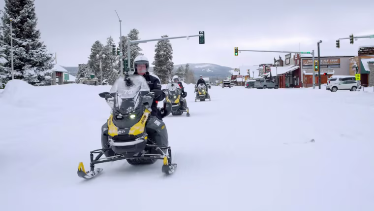 A group of people driving snowmobiles in the snow on the street outside
