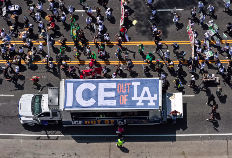 A aerial view of a truck with a banner on top that reads "ICE out of LA", driving next to a large group protestors walking down the street