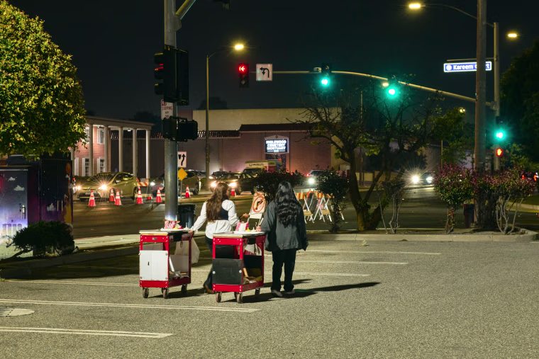 Two women walk outside rolling food carts beside them in a parking lot next to a street, their backs are to camera