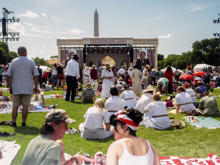 People sit on the grass and stand in front of an outdoor stage with the Washington Monument visible in the background.
