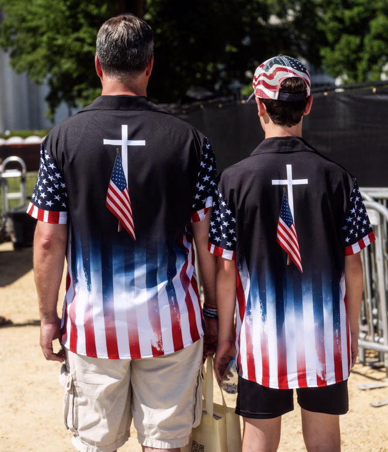 A father and son stand with their backs to the camera wearing polo shirts with a white cross overlayed by the American flag.