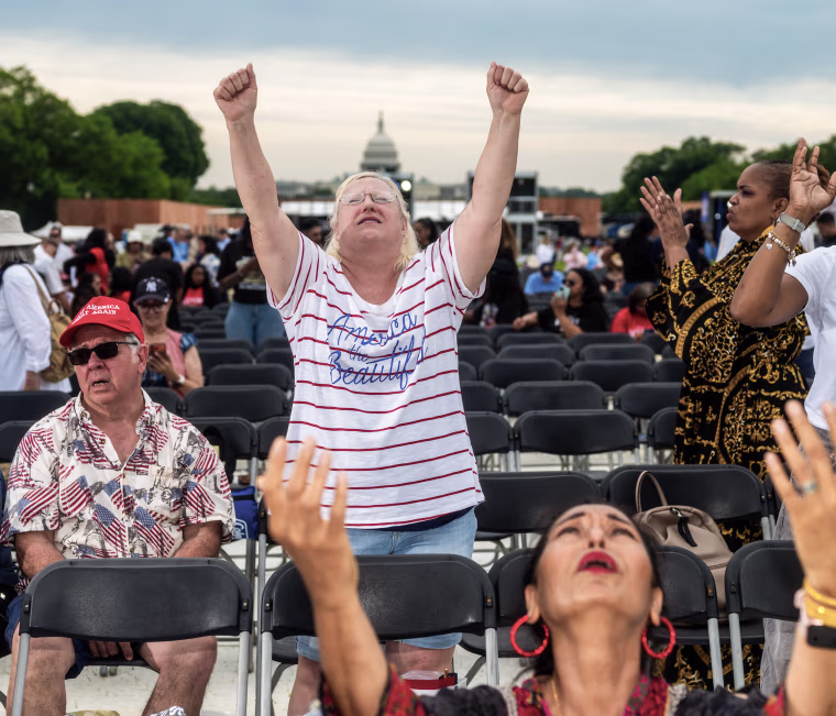 Two women raise their hands to the sky in prayer while others sit nearby on rows of folding chairs with the U.S. Capitol building visible in the background