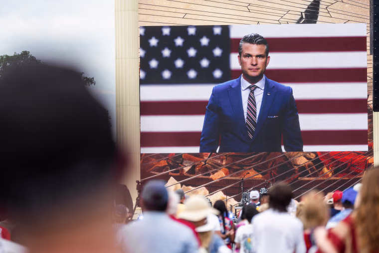 Pete Hegseth appears on a jumbo screen in front of an outdoor audience.