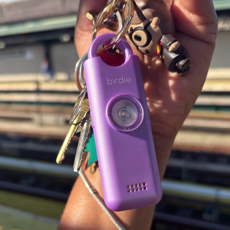 The Birdie personal alarm on a keychain, behind held up in front of outdoor subway tracks.