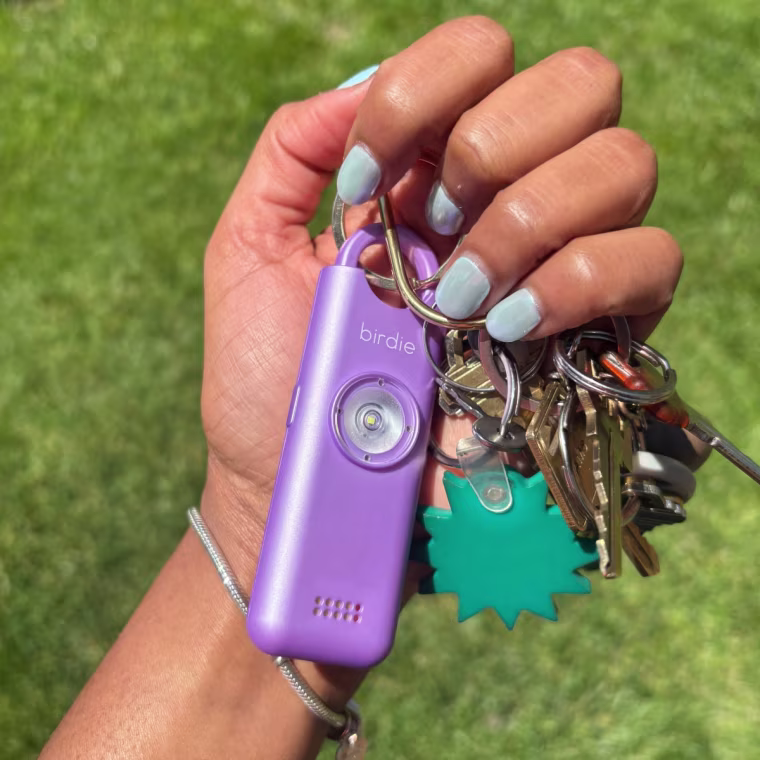 The Birdie safety alarm on a keychain while being held in a hand with blue fingernails against a grass background.