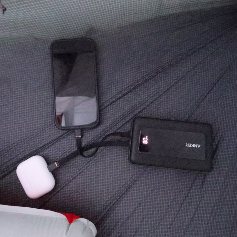 A power bank charging an iPhone and Apple AirPods on the floor of a tent.