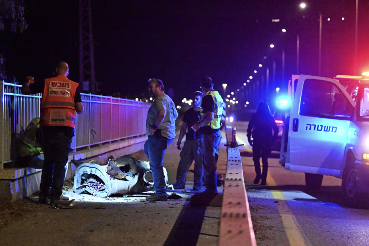Emergency responders stand next to charred remnants of a rocket on a roadway.