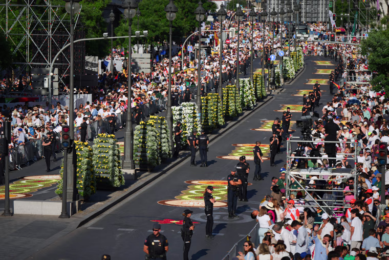 Flower petal beds can be seen on the street in between rows of people crowded on the sidewalks