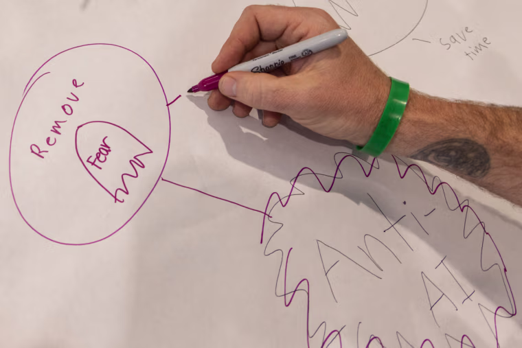 A hand uses a sharpie to write "Anti-AI, remove fear" in magenta on a piece of large white paper