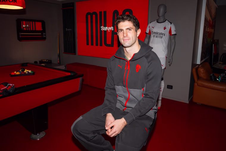 Christian Pulisic sits on a chair next to a pool table inside of a red-themed sports room