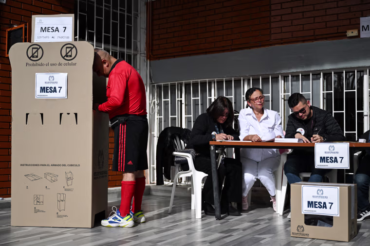 A man votes at a polling station booth next to a table of poll workers inside of a room