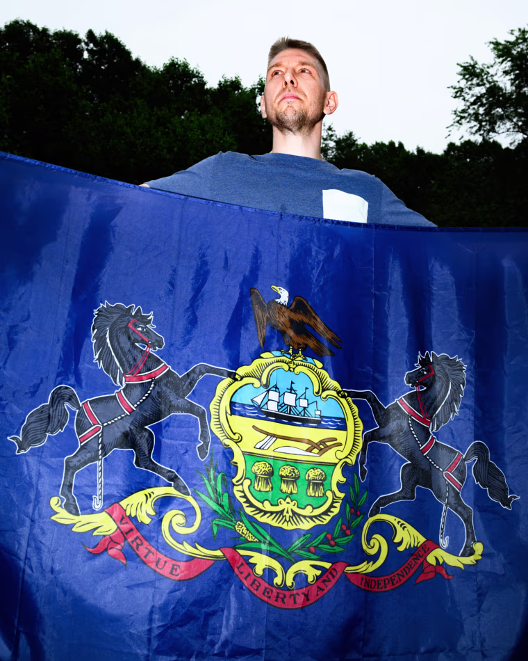 Frank Chappell stands outside holding a blue flag with an insignia showing two horses, an eagle, and a ship in the center crest, text on the bottom on a red ribbon design reads "virtue, liberty, and indepence"