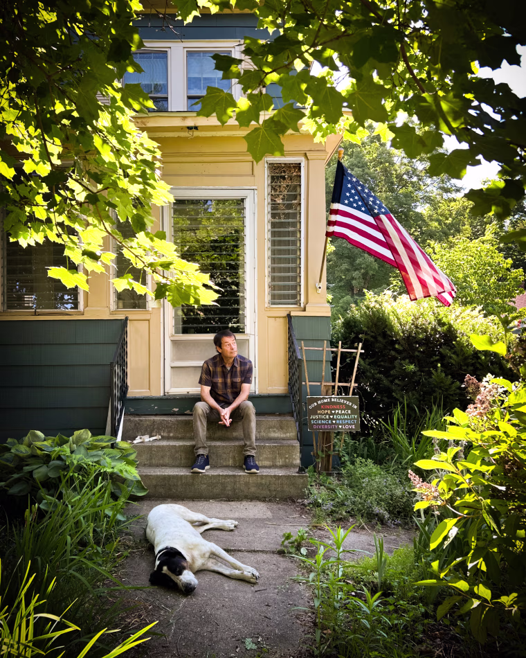 Bruce Watson sits on the front steps to his house, his dog lays on the path in front of him, and an American flag flies in front of the house
