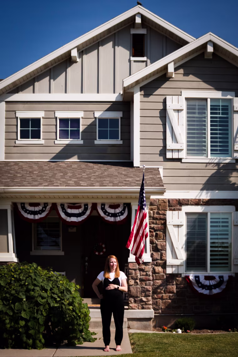 Rebecca Dyer, stands in front of her home with a hand on her pregnant belly, an American flag can be seen flying in front of the house