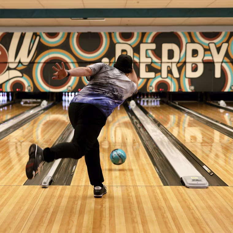 A man kicks one leg back behind him after throwing a bowling ball down a lane.