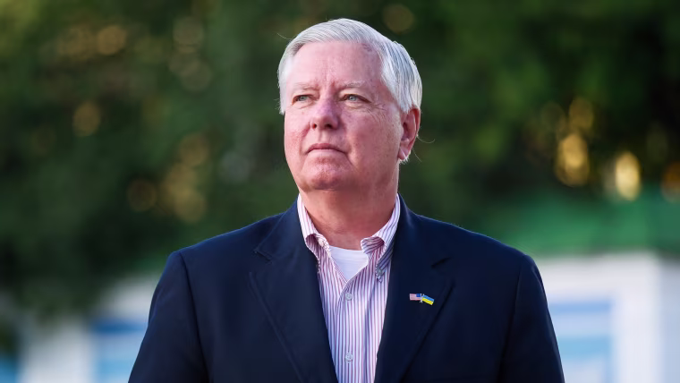 Lindsey Graham stands outside and looks off in the distance, he wears a pin on his dark blue suit's lapel that shows the U.S. flag and Ukraine flag side by side