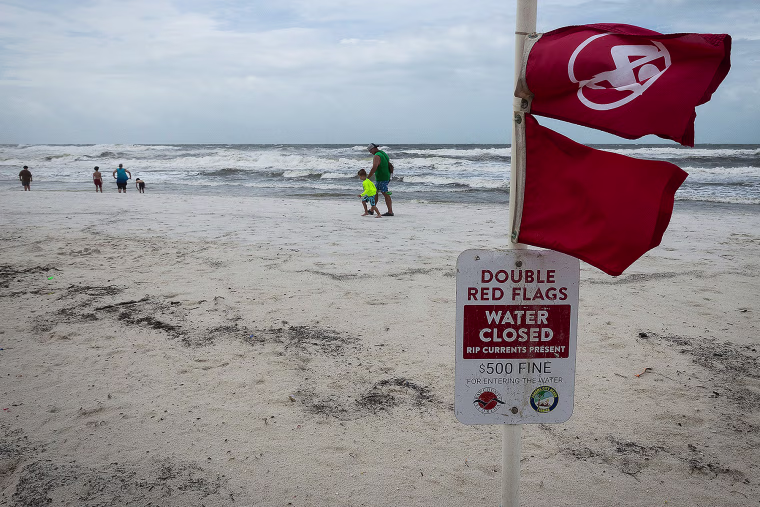 People walk by the shore on the beach, in the foreground are two red flags on a pole above a sign that reads "Double Red Flags, Water Closed, Rip Currents Present, $500 Fine for entering the water"