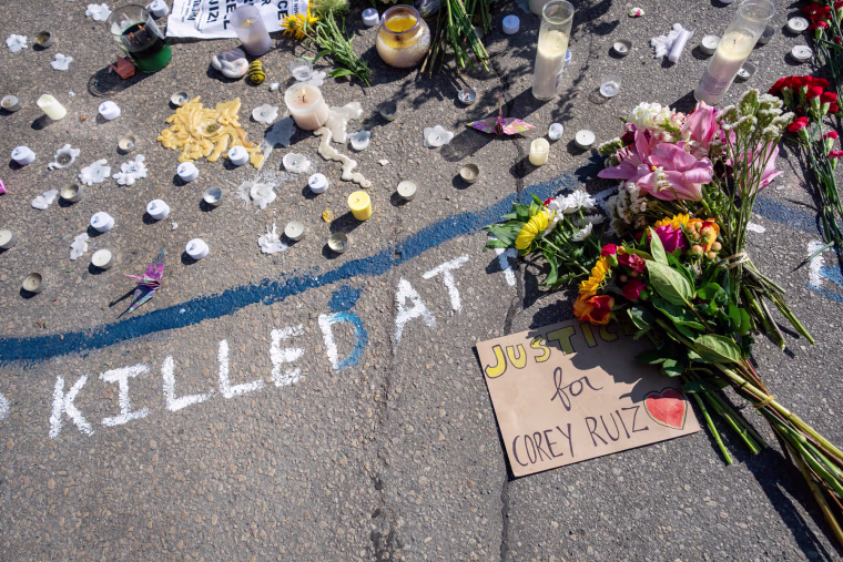 Candles, flowers, and a cardboard sign saying "justice for Corey Ruiz" are seen on a street. Written in walk chalk next to the poster reads "Killed at..."
