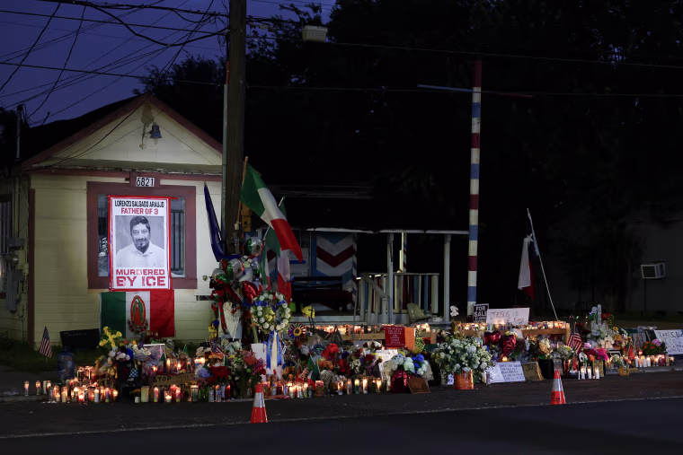 A makeshift memorial with dozens of candles and flowers laid in front of a small house at night.