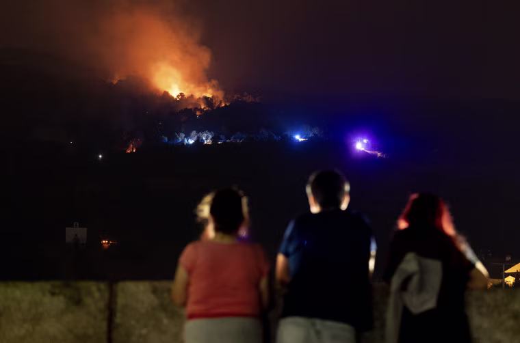 Three people are silhouetted as they face toward a distant wildfire at night.
