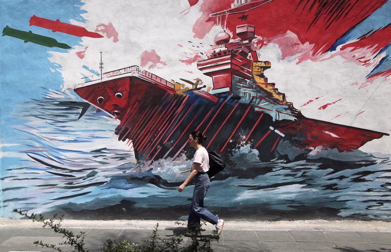 A young adult wearing earbuds walks in front of a war propaganda mural painted onto the side of a building.
