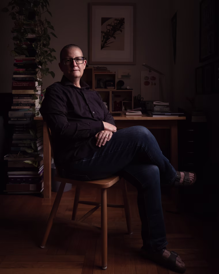 A middle-age white woman with a shaved head sits with crossed legs in front of her desk and large stacks of books.