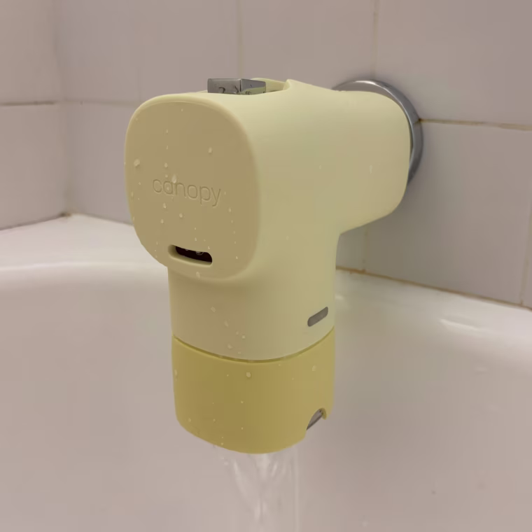 A yellow Canopy Bath Tub Filter installed on a tub spout against a white tile background.