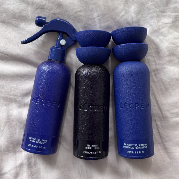 The Cecred Detangling Spray, Oil Ritual and Detoxifying Shampoo lined up on a light pink bedspread.