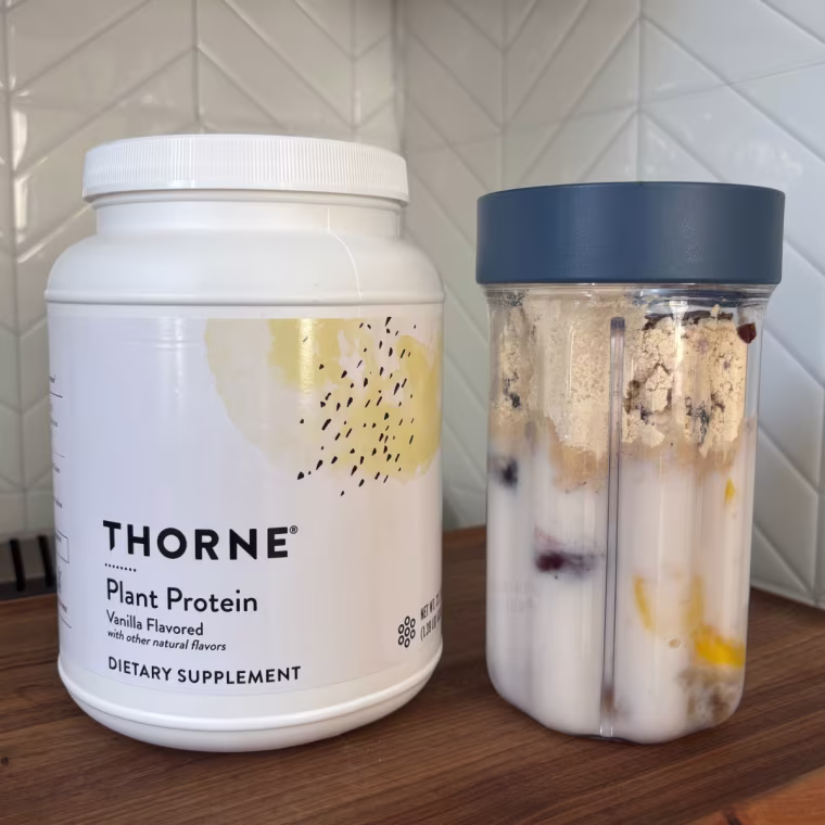 A container of Thorne’s Vanilla Plant Protein next to a smoothie in a clear plastic cup with a straw.