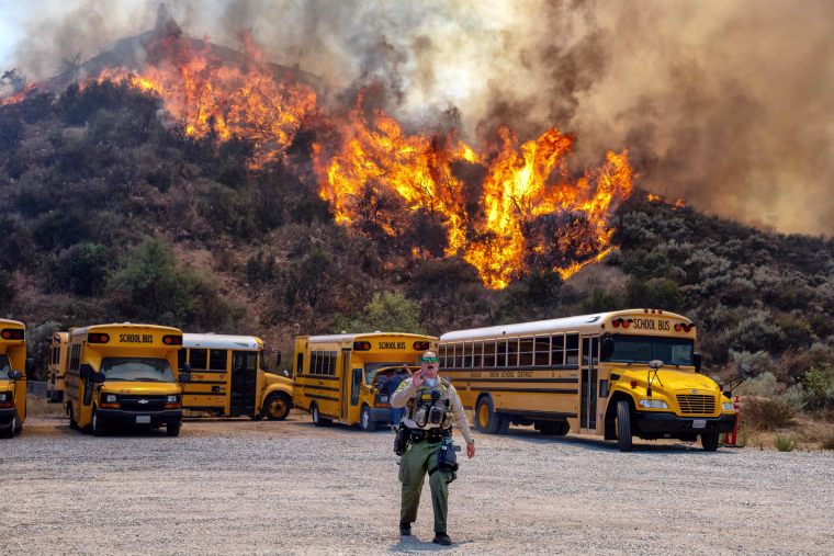 A brush fire is seen in the trees behind a row of parked school buses in a parking lot as a cop walks in the foreground speaking